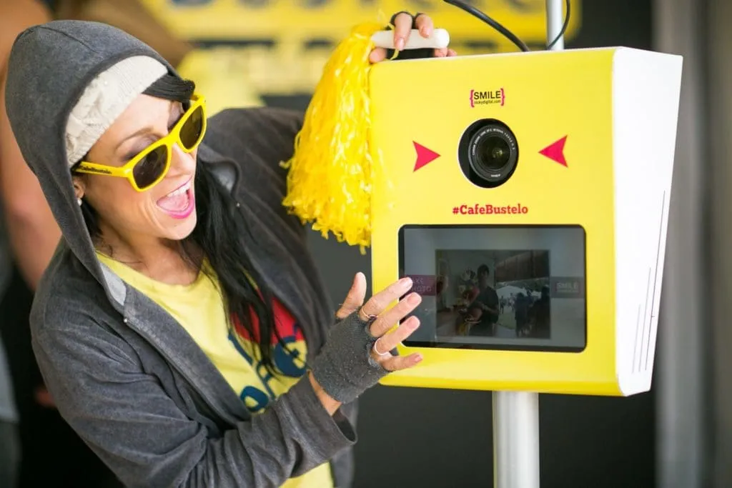 Guest in yellow sunglasses and black hoodie smiling at a bright yellow AI photo booth branded with #CafeBustelo at an event