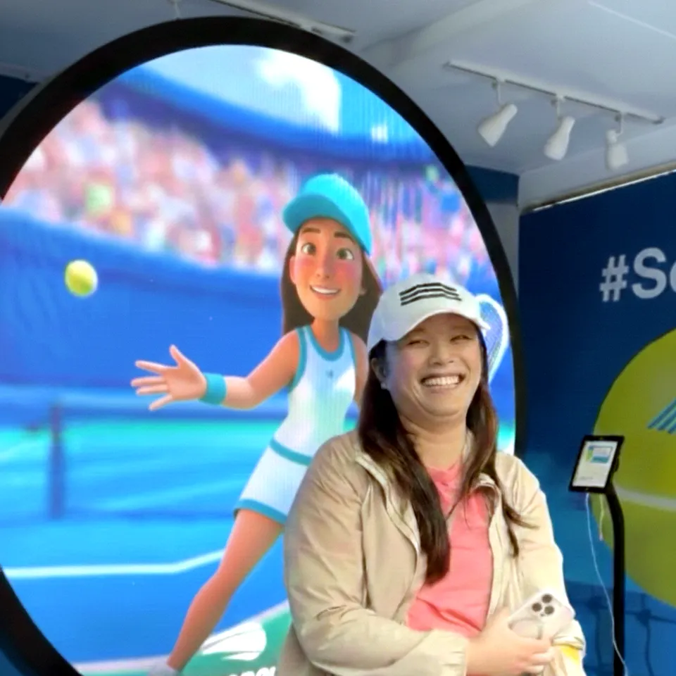 Smiling woman in white cap and pink shirt posing in front of circular display screen showing AI-generated avatar of tennis player in blue outfit on court