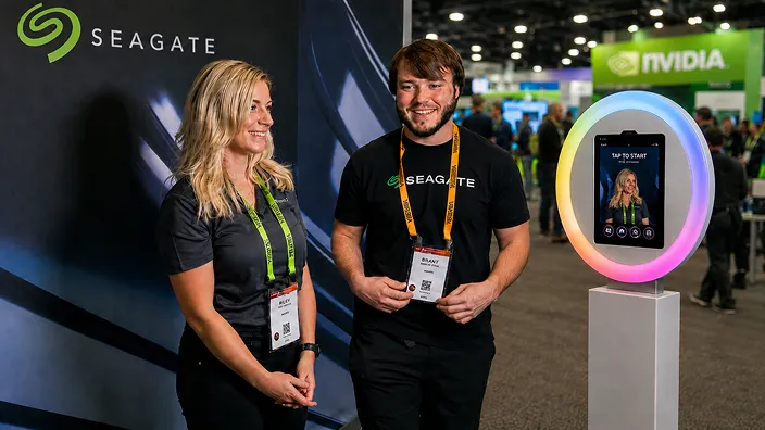 Two trade show attendees wearing lanyards stand beside an AI photo booth with illuminated circular frame displaying a selfie interface at a tech conference.
