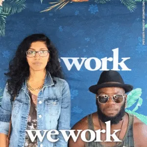 Two people posing in front of a blue WeWork branded photo backdrop with tropical leaf graphics