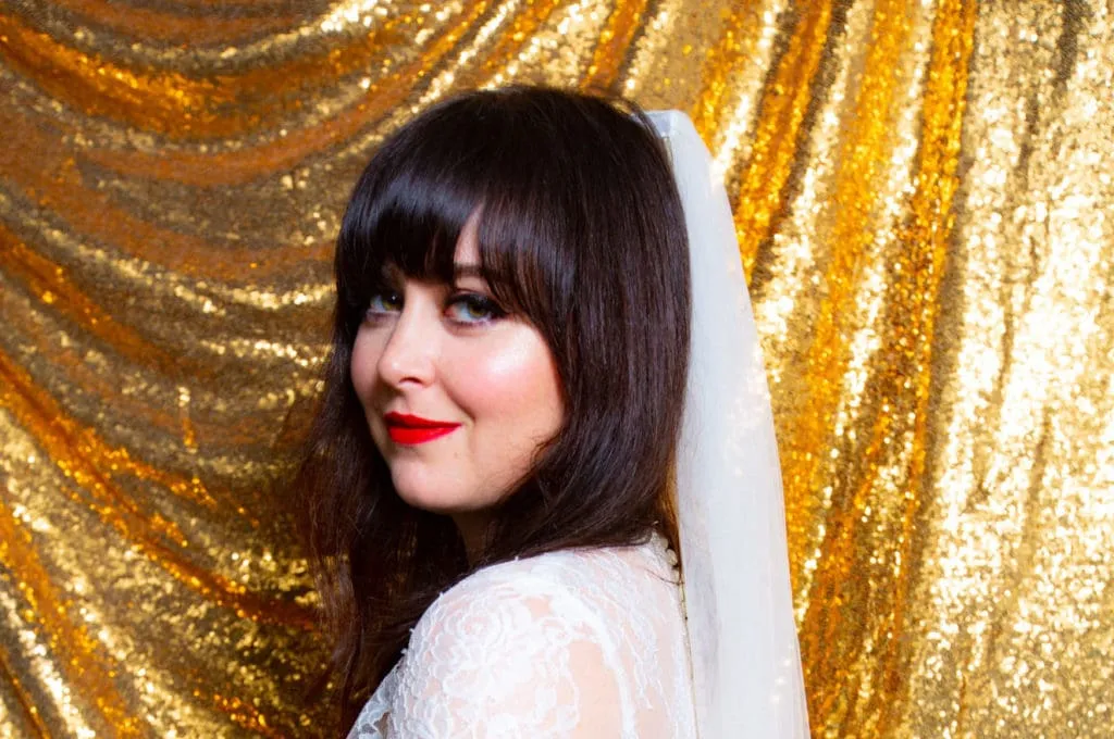 Bride in white lace veil and red lipstick poses in profile against gold sequin backdrop at wedding photo booth