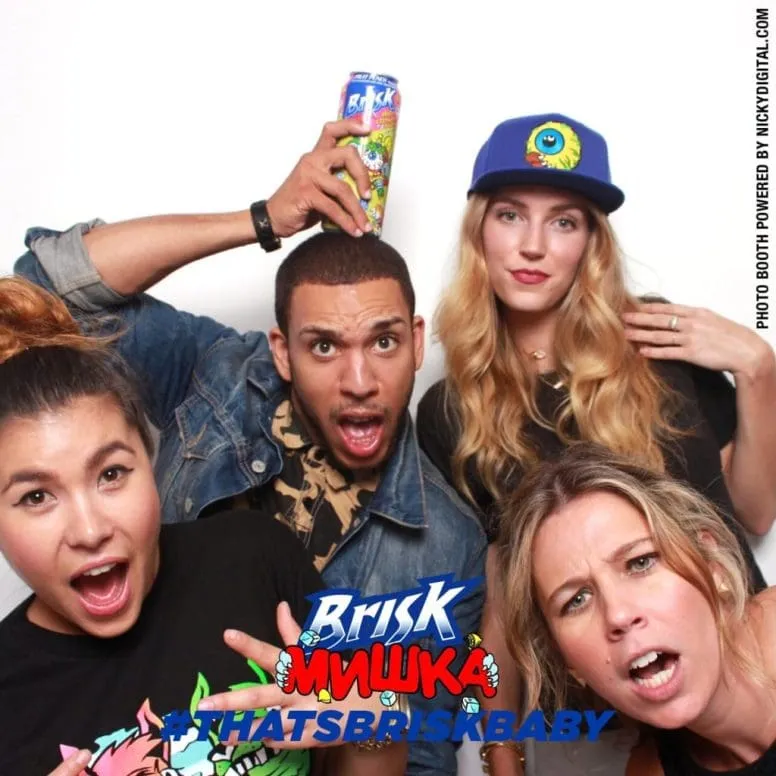 Four people posing together with a Brisk-branded photo booth prop displaying playful expressions and holding a colorful beverage can.