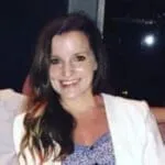 Woman in white blazer over blue top smiling at camera against dark background
