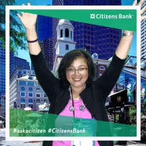 Woman with arms raised in celebration at Citizens Bank trade show photo booth, wearing pink top and glasses, with city skyline backdrop
