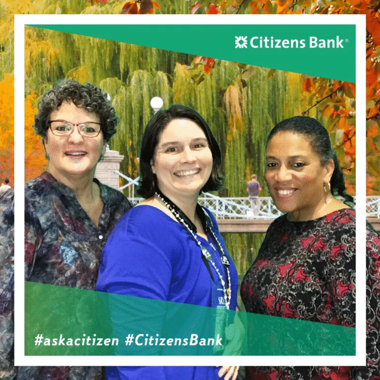 Three smiling women posing together at Citizens Bank trade show booth with autumn foliage backdrop