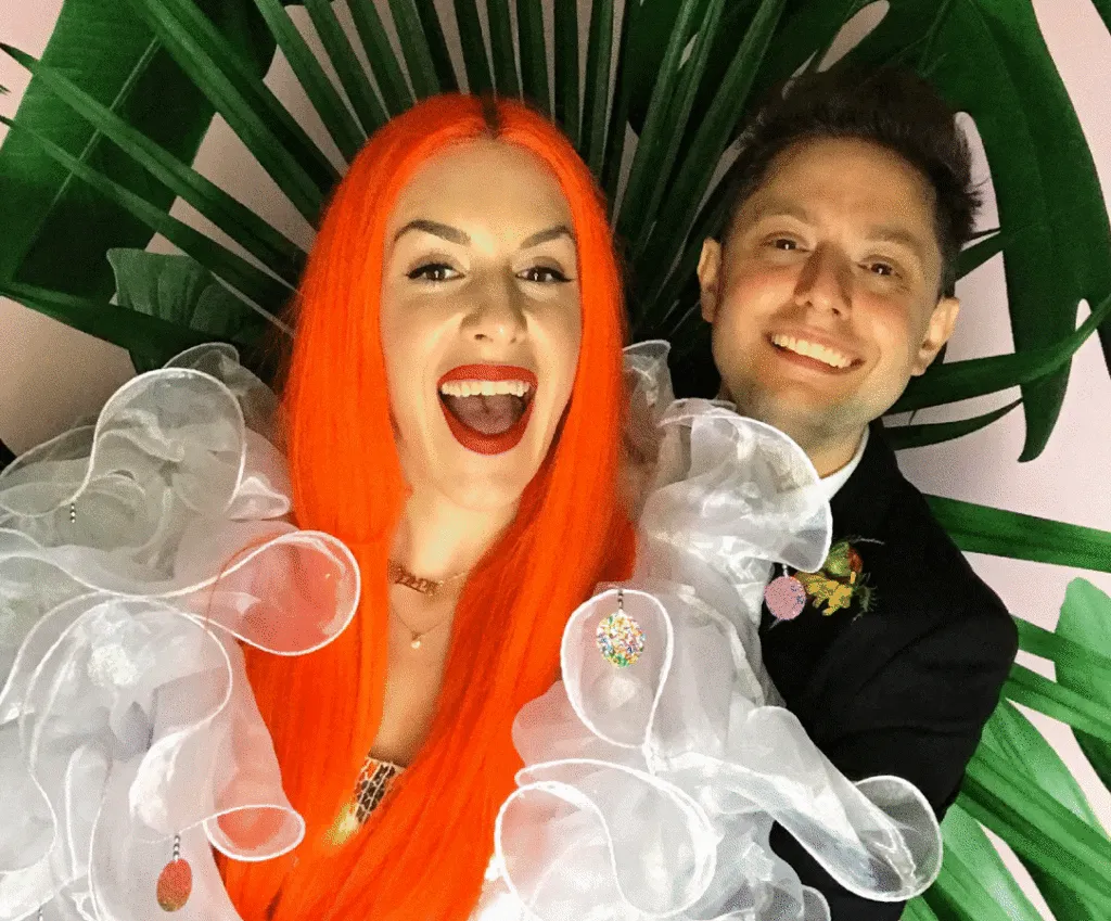 Couple smiling at open-air photo booth with tropical leaf backdrop, woman wearing bright orange wig and sheer white ruffled shoulders, man in dark blazer