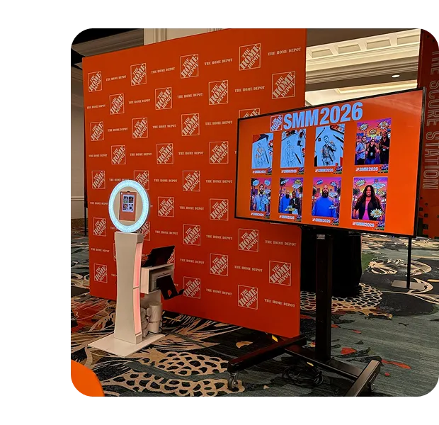 SIMM2026 branded photo booth experience OutSnapped FutureFoto photo booth with white pedestal kiosk at SMM2026 brand activation, branded orange backdrop with repeated logo pattern and digital display showing guest photos