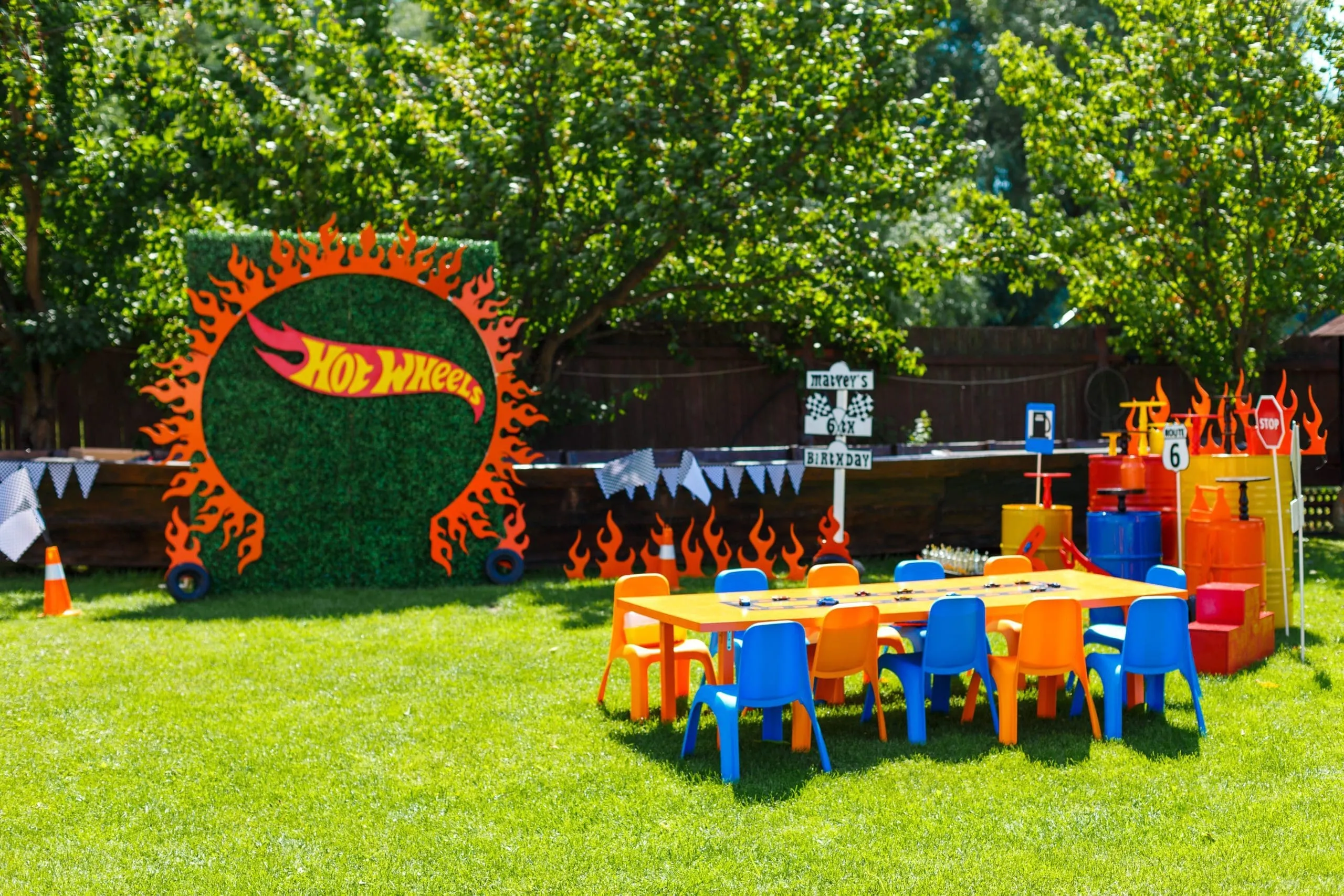 Hot Wheels Racing Birthday Party Backdrop Hot Wheels-themed outdoor party setup with flame backdrop, colorful children's table and chairs on grass, and branded signage.