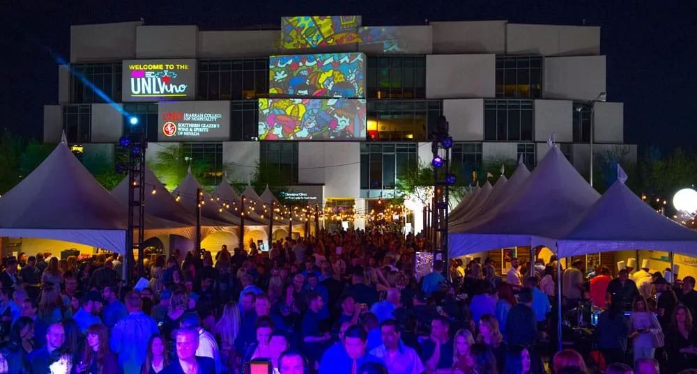 Large outdoor nighttime event at Keep the Memory Alive Event Center in Las Vegas, with purple-lit tent structures, crowd of attendees, and illuminated modern building facade in background