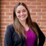 Kira Holt - OutSnapped Customer Testimonial Woman smiling at camera wearing navy cardigan and purple top against red brick wall