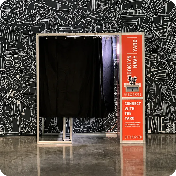 MagBooth enclosed photo booth with black curtain and cream frame, branded signage for Brooklyn Navy Yard behind monochrome doodle backdrop.