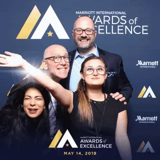 Branded Corporate Awards Photo Booth Backdrop Four people posing at Marriott International Awards of Excellence event with branded backdrop featuring gold and blue geometric logo, May 14, 2018.