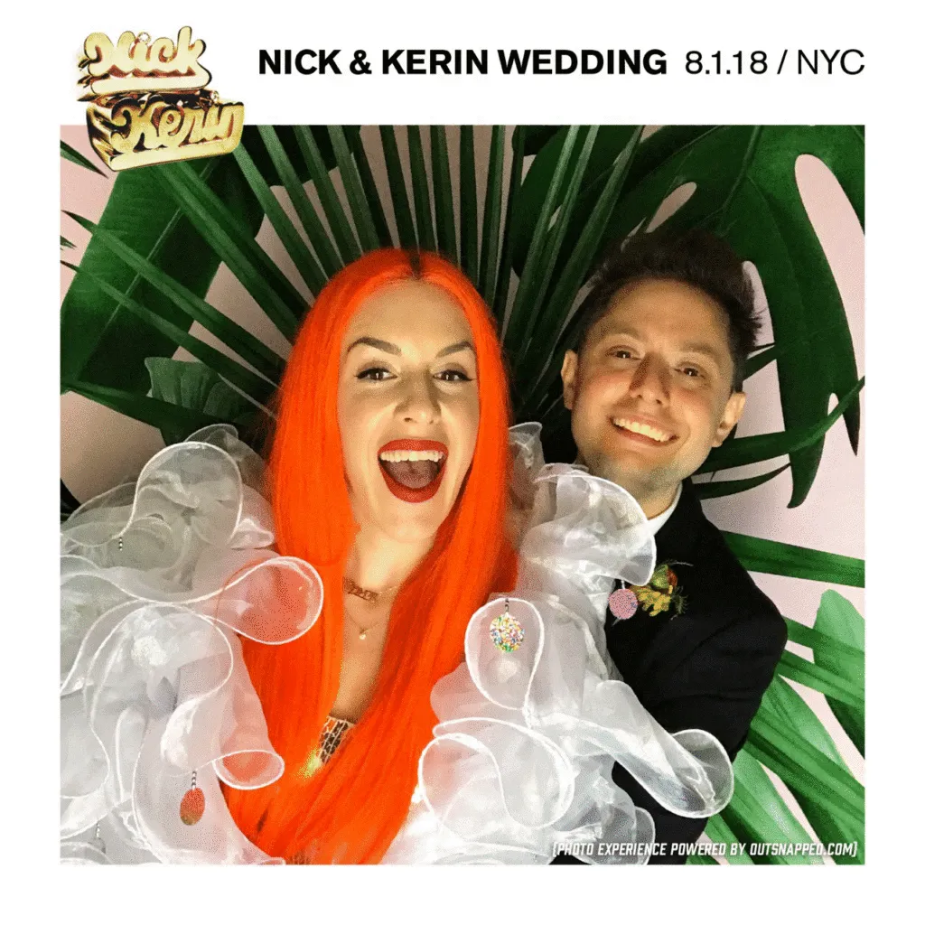 Bride with bright orange hair in white ruffled gown and groom in black tuxedo smile at NYC wedding photo booth, tropical leaf backdrop behind them