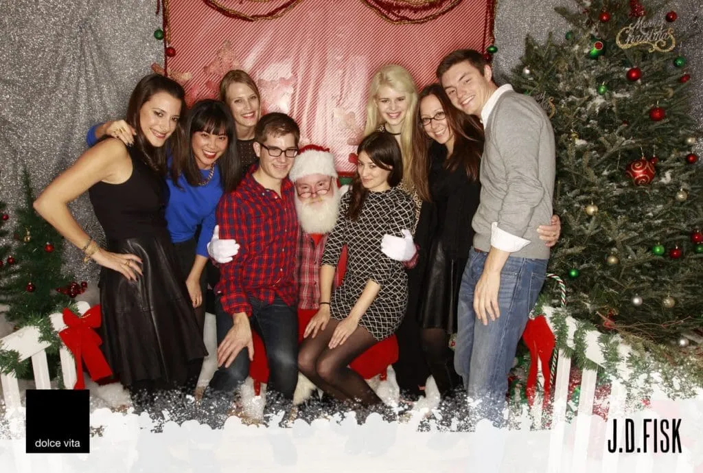 Group of eight people smiling at an office holiday party photo booth, dressed festively with Santa hat and holiday attire, posed in front of decorated Christmas tree and pink backdrop with fake snow