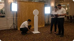 Premium 360 Photo Booth Installation Team members at a corporate event demonstrating an open-air photo booth setup with wooden backdrop and professional lighting