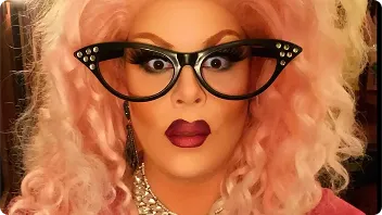 Person wearing cat-eye glasses and pink wavy wig with bold red lipstick, smiling at camera