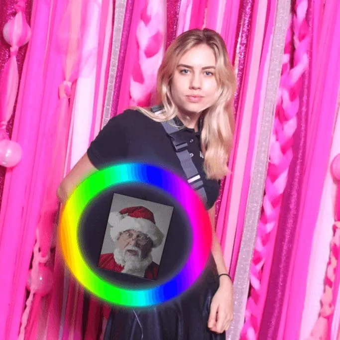 Woman posing in front of vibrant pink and silver backdrop with colorful ring light creating neon green, blue, and yellow glow effect around instant photo display