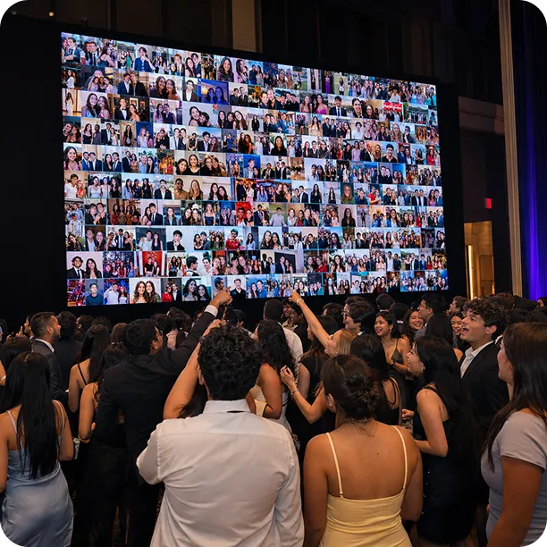Crowd of formally dressed guests viewing a large digital display wall showing a grid of hundreds of photo booth snapshots from a university event.
