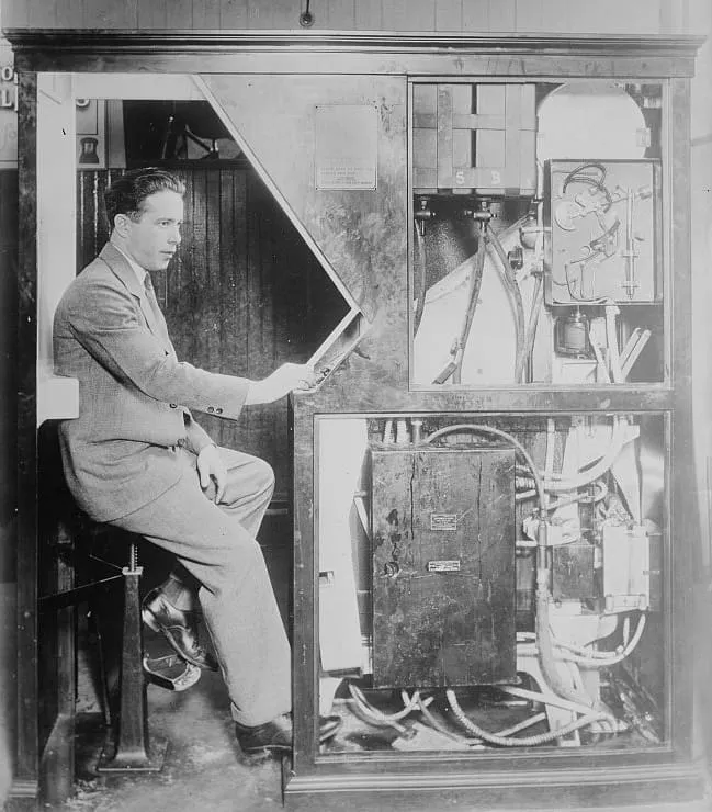 Man in suit operating vintage photo booth machinery from the 1920s-1930s, examining internal mechanical and electrical components through open cabinet panels.