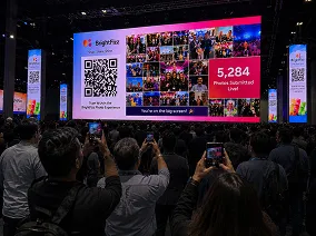 Real-Time Photo Wall at Large-Scale Event Crowd of event attendees viewing large projection screen displaying virtual photo booth gallery with QR code and photo count at trade show or corporate event