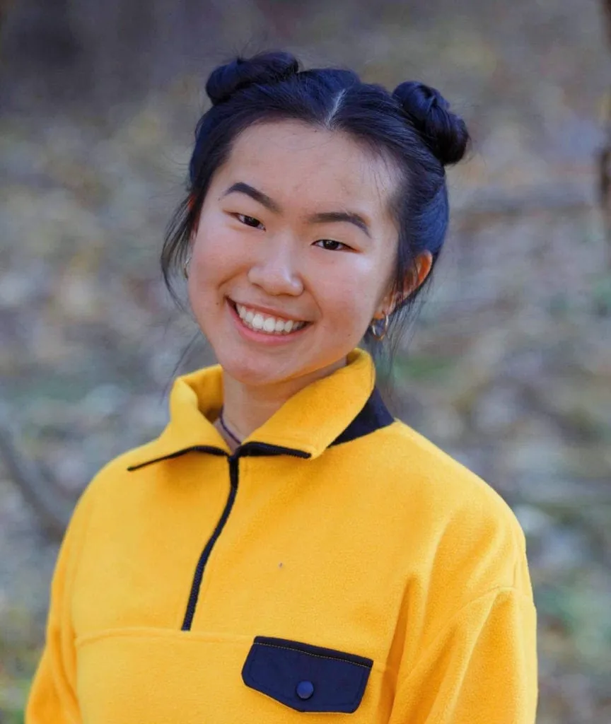 Headshot of Zimmy Kang smiling in a yellow fleece with space-bun hair, photographed outdoors