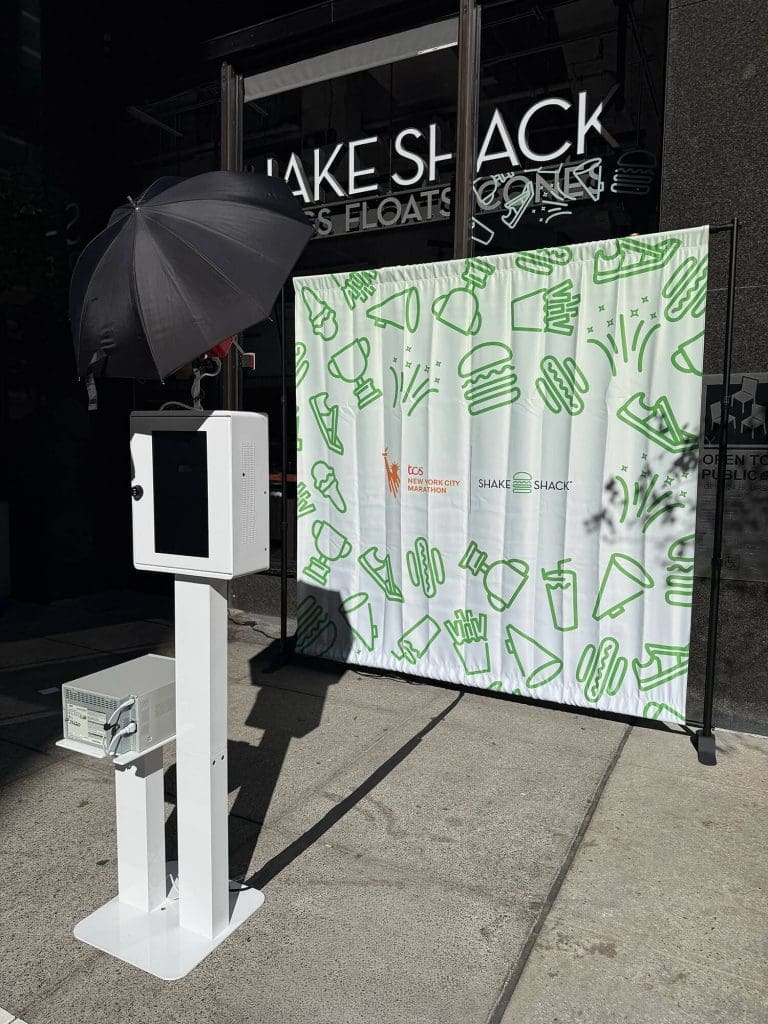 NYC Marathon Photo Booth Rental Case Study: How Shake Shack Generated 50K+ Impressions at the Finish Line 1 OutSnapped FutureFoto open-air photo booth rental setup with professional strobe lighting and instant flash at NYC Marathon, featuring branded Shake Shack and TCS Marathon backdrops with green pattern design
